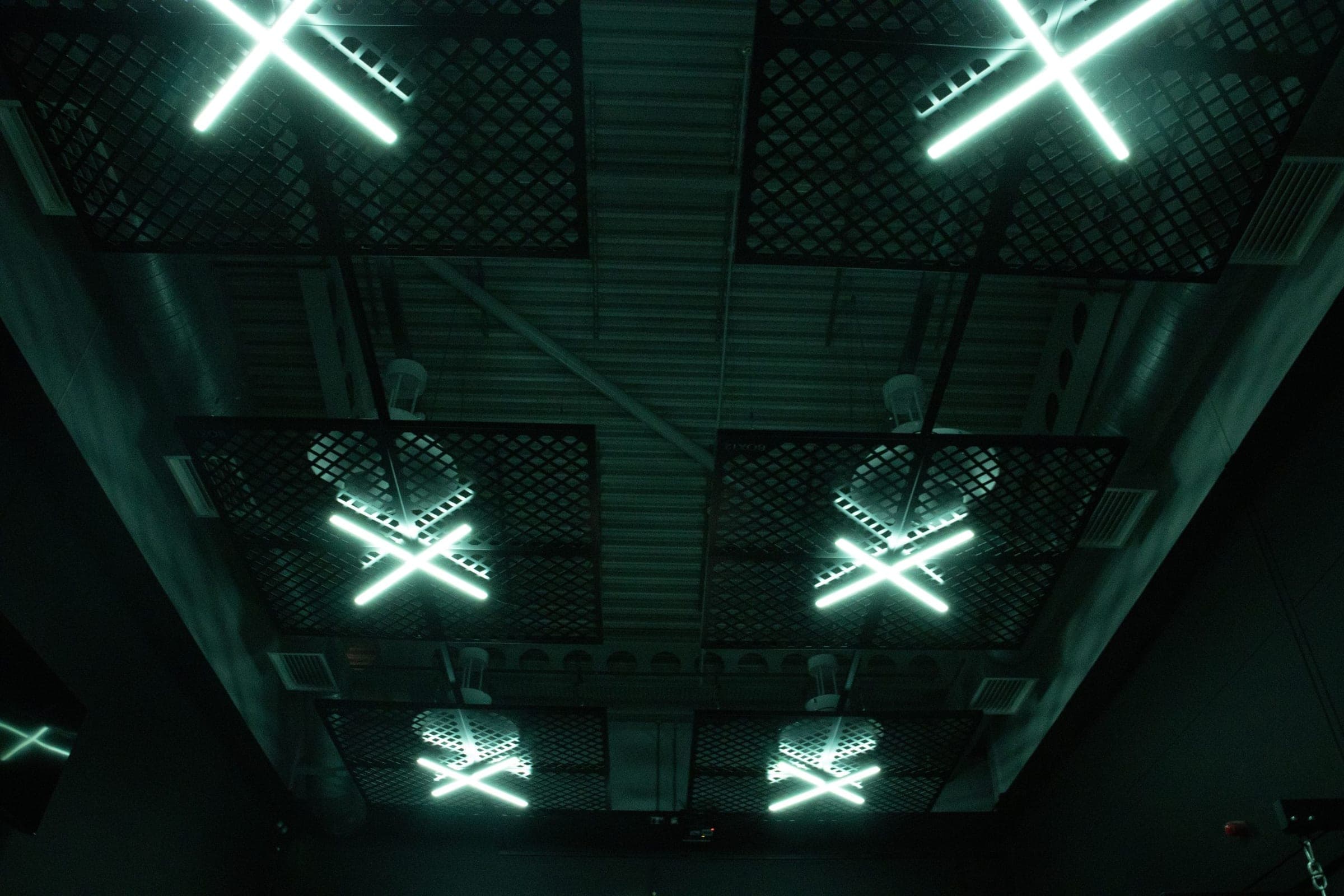 Cross-shaped teal neon tube lights on a dark industrial ceiling mesh grid, BOX12