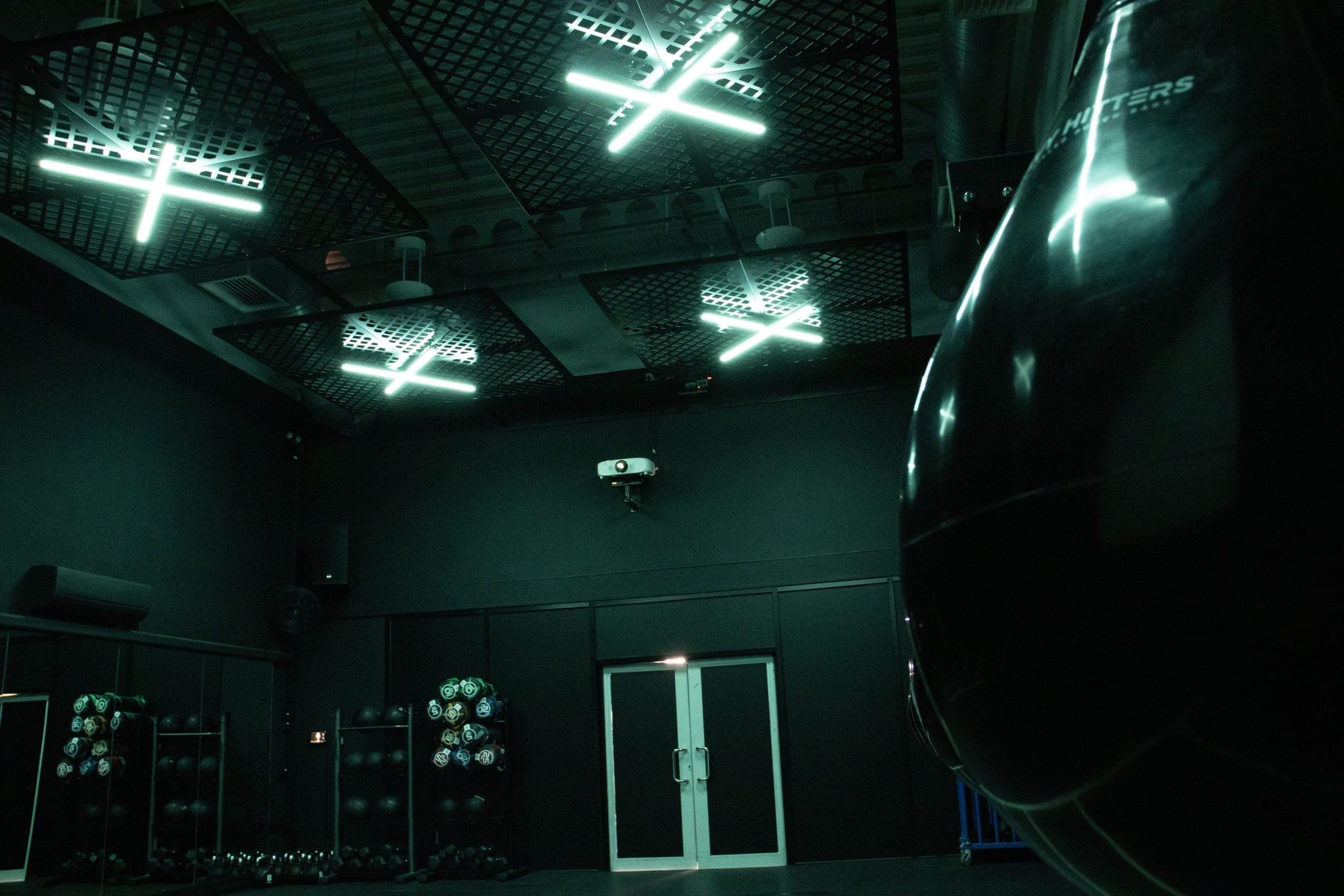 BOX12 gym interior in teal-green lighting with heavy bag foreground and kettlebells racked along the wall
