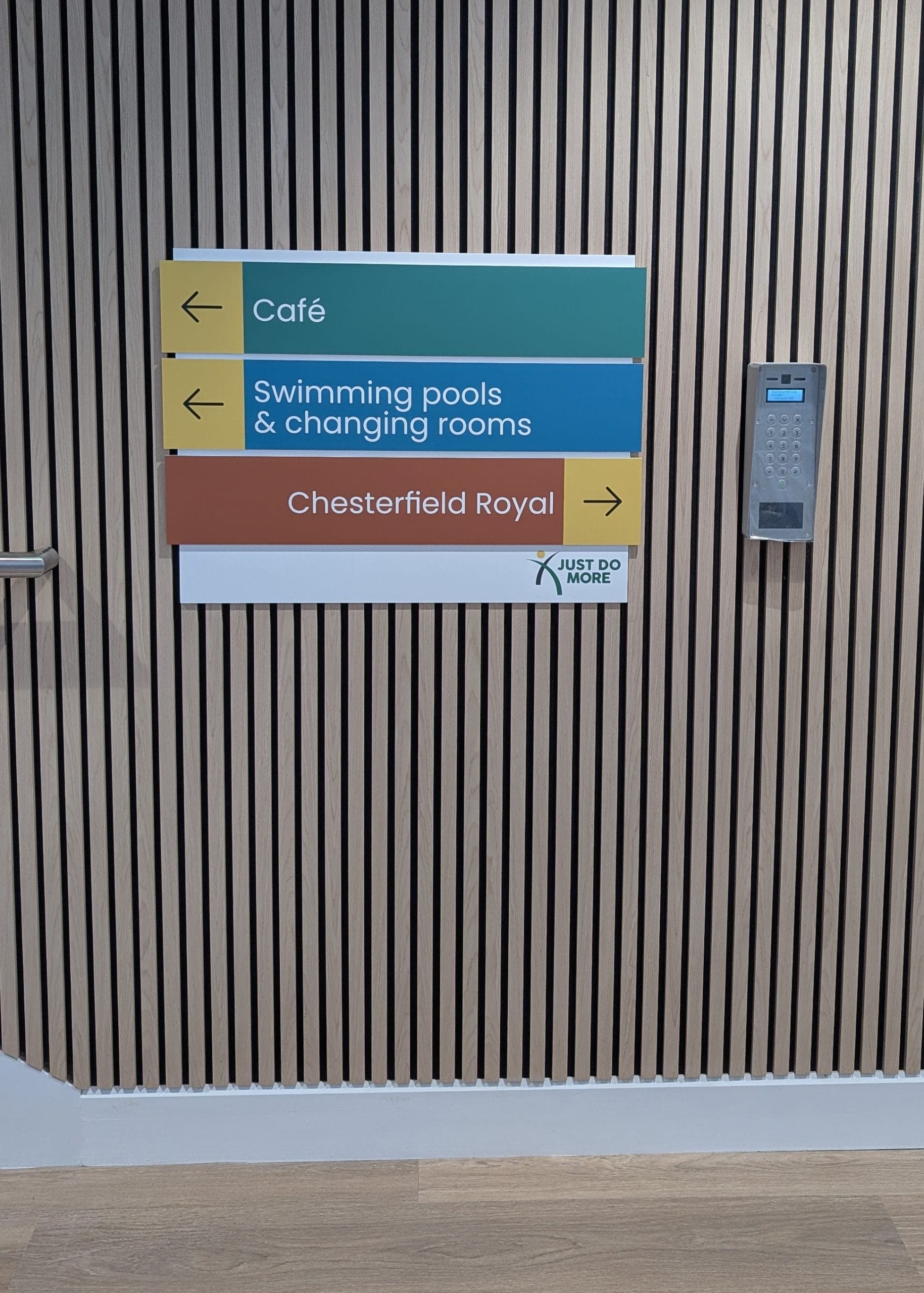 Multi-colour directional wayfinding sign panel on slatted wood wall pointing to café, pools and Chesterfield Royal