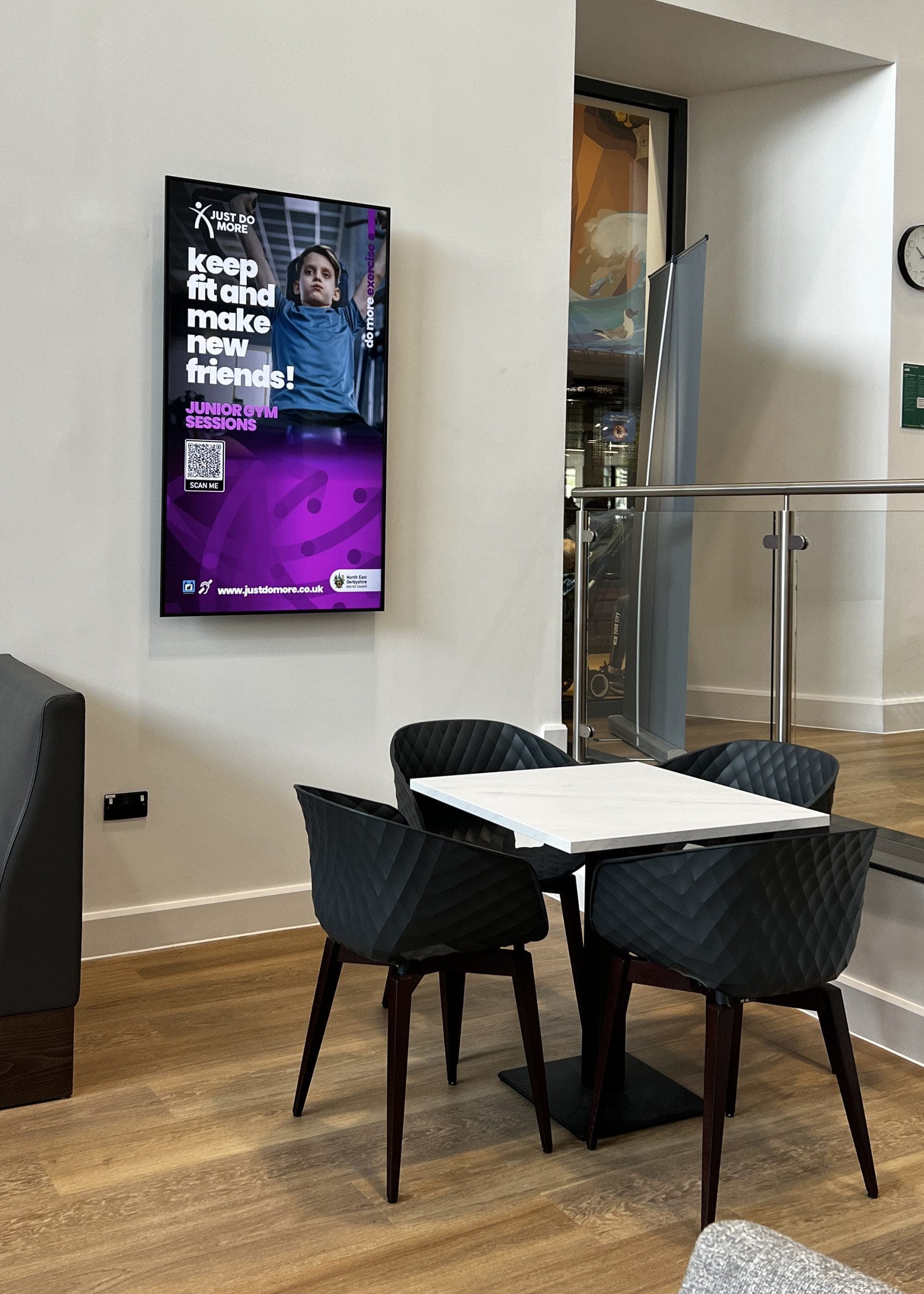 Café seating area with wall-mounted portrait digital screen showing Junior Gym Sessions advert