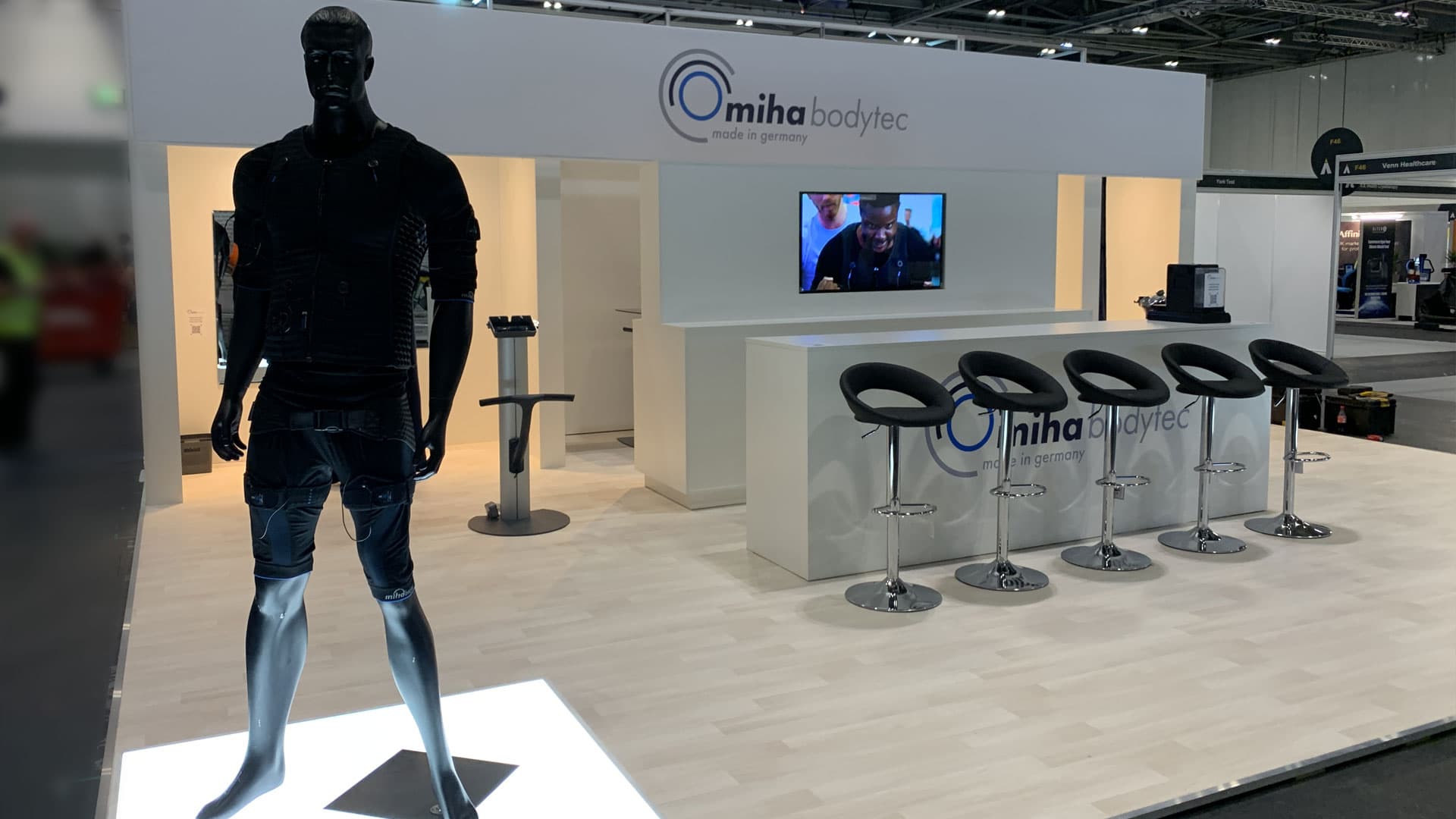 Full miha bodytec stand with black mannequin in EMS suit, branded reception counter and bar stools