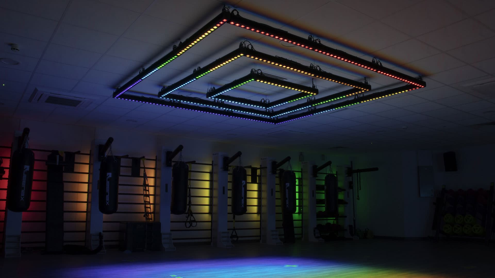 Concentric-square ceiling rig lit in full rainbow spectrum casting coloured pools on the gym floor